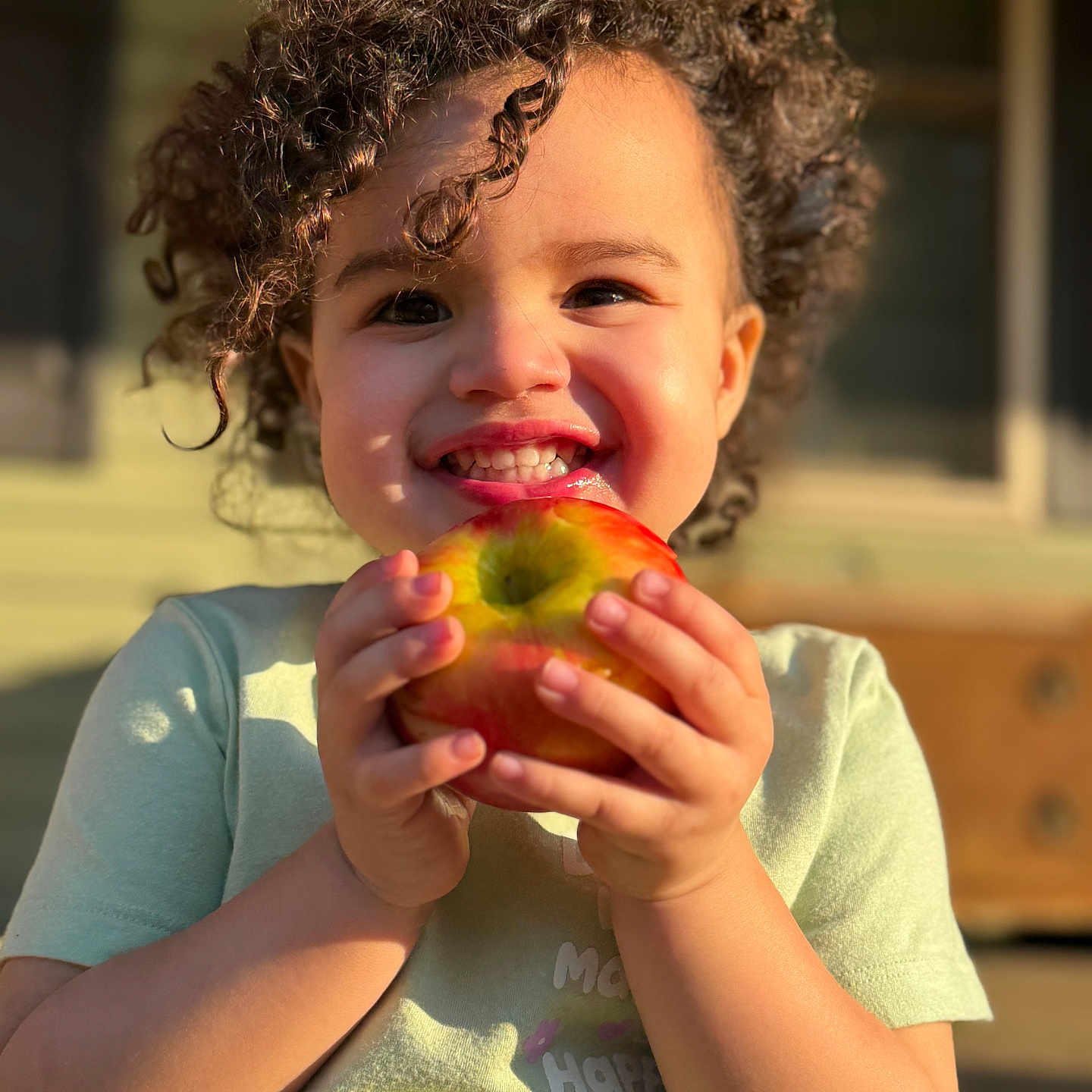 Kyah joined the competition — help win amazing prizes! apple, cheerful, child, close_up, curly_hair, cute, face, happy, holding, natural_light, outdoor, person, playful, portrait, short_sleeve_shirt, skin, smiling, sunlight, toddler, young_child