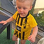 toddler, child, yellow_jersey, soccer_shirt, standing, balcony, outdoor, wicker_table, hand, curly_hair, shoe, green_carpet, wooden_floor, window, reflection, building, daylight, person, casual_clothing, snack