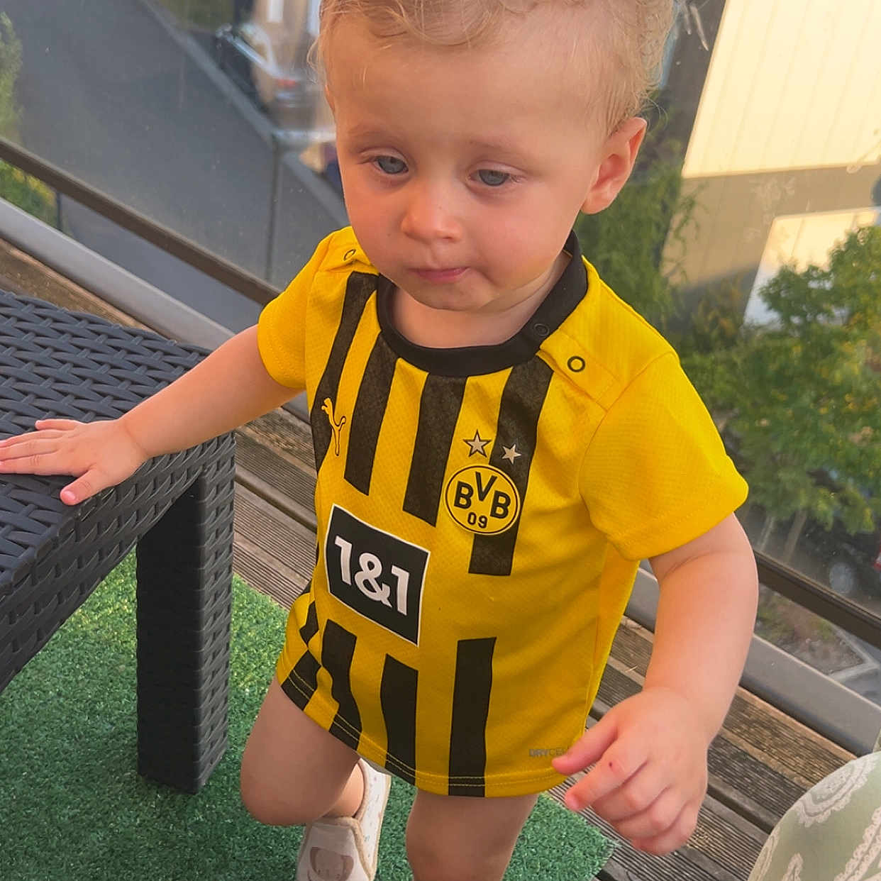 Tiago a rejoint le concours — aidez-le/la à gagner de superbes lots ! balcony, building, casual_clothing, child, curly_hair, daylight, green_carpet, hand, outdoor, person, reflection, shoe, snack, soccer_shirt, standing, toddler, wicker_table, window, wooden_floor, yellow_jersey