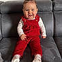 baby, child, comfortable, corduroy, couch, cute, floral_pattern, gray_couch, hair_tufts, happy, indoor, infant, long_sleeve_shirt, person, portrait, red_overalls, sitting, smiling, socks, young_child