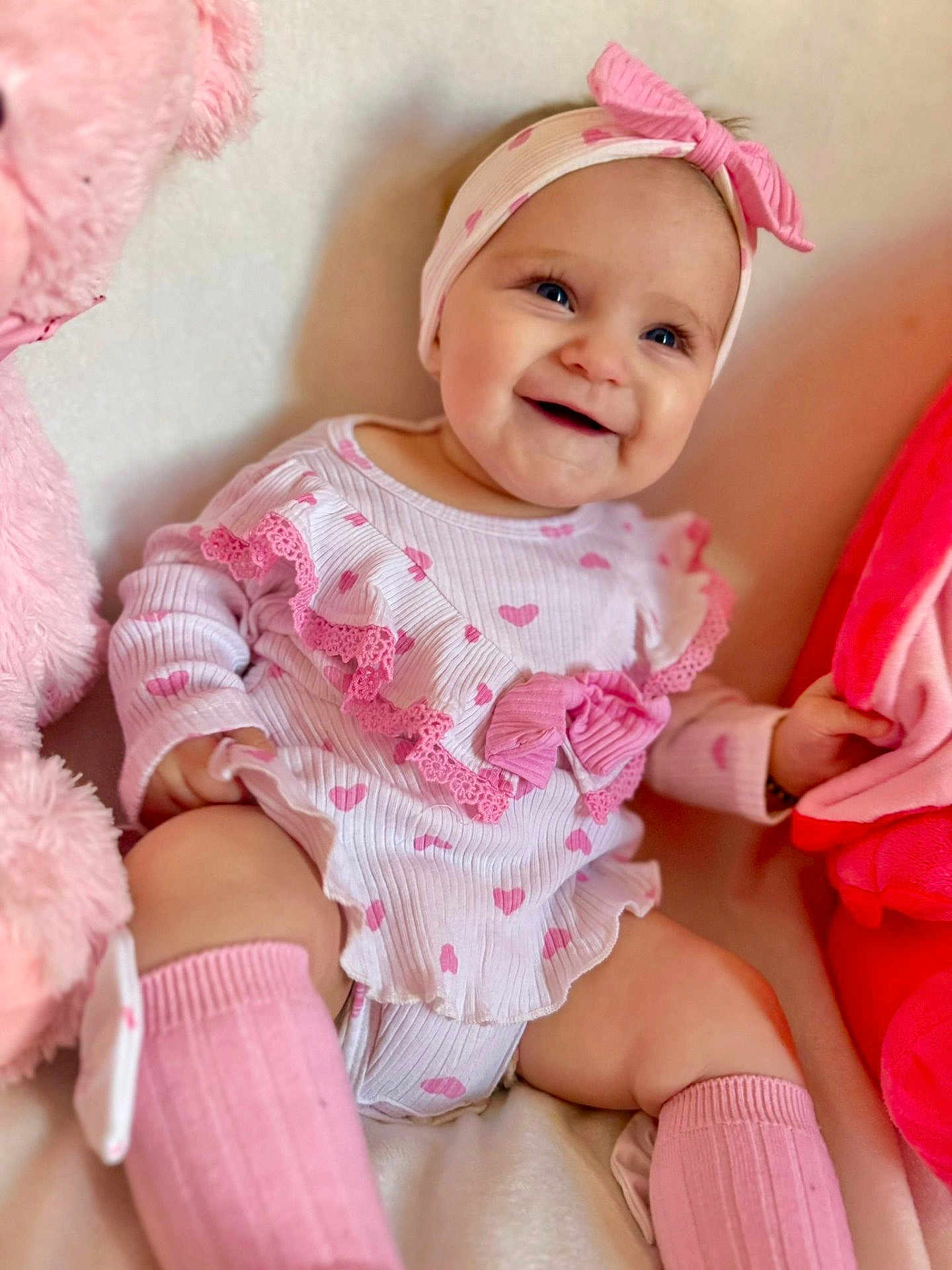 Aria is registered to the contest to win money with this photo: baby, infant, smiling, pink, headband, heart_pattern, sitting, plush_toy, soft_toys, cute, child, clothing, sock, happy, portrait, indoors, comfort, cute_expression, baby_girl, warm_colors