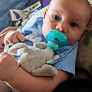 David is registered to the contest to win money with this photo: baby, pacifier, plush_toy, blue_shirt, closeup, child, infant, soft_toy, hand, face, eyes, cute, indoors, comfort, sleepy, cuddling, person, childhood, relaxation, portrait