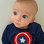 baby, child, infant, face, blue_eyes, sweater, captain_america, superhero_logo, curious_expression, portrait, close_up, cute, young_child, clothing, casual_wear, indoor, light_background, head, person, looking_away