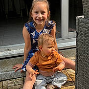 Layana a rejoint le concours — aidez-le/la à gagner de superbes lots ! child, toddler, smile, outdoor, sunlight, wooden_deck, wicker_furniture, dress, casual_clothing, happy, sitting, family, portrait, person, baby, floor, door, bracelet, earrings, hair_tie