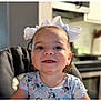toddler, child, smiling, curly_hair, white_bows, high_chair, blue_shirt, cartoon_print, indoor, kitchen, happy, person, face, cute, young_child, portrait, seated, domestic, background_blur, home