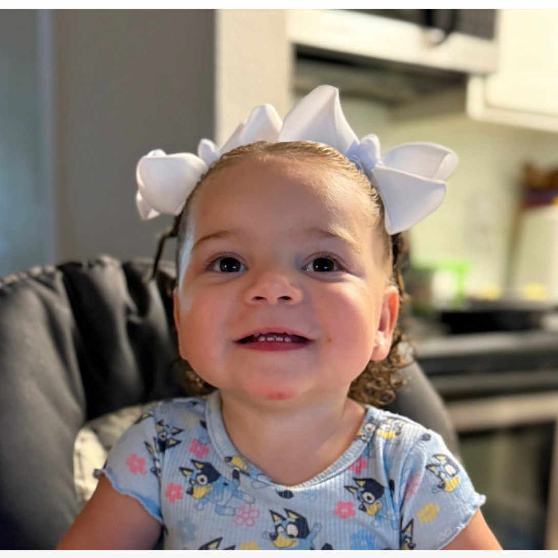 Gema Valentina joined the competition — help win amazing prizes! background_blur, blue_shirt, cartoon_print, child, curly_hair, cute, domestic, face, happy, high_chair, home, indoor, kitchen, person, portrait, seated, smiling, toddler, white_bows, young_child