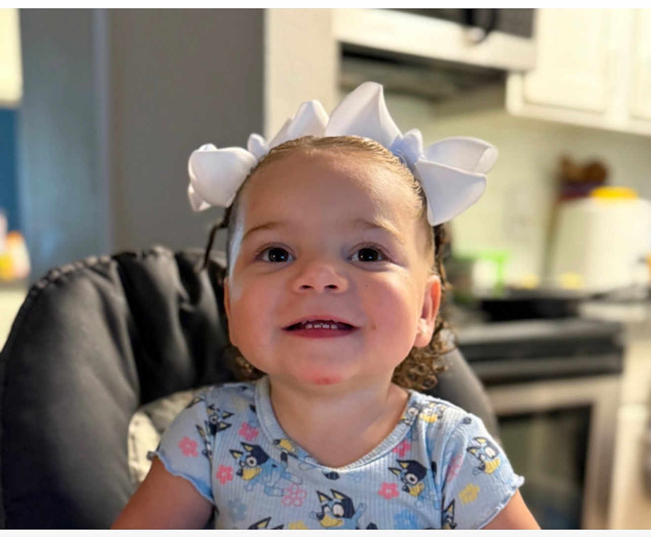 Gema Valentina joined the competition — help win amazing prizes! toddler, child, smiling, curly_hair, white_bows, high_chair, blue_shirt, cartoon_print, indoor, kitchen, happy, person, face, cute, young_child, portrait, seated, domestic, background_blur, home