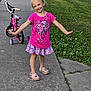 child, girl, smiling, sidewalk, bicycle, training_wheels, pink_shirt, floral_skirt, shoes, grass, outdoor, playful, happy, posing, daylight, fence, concrete, person, casual_clothing, nature