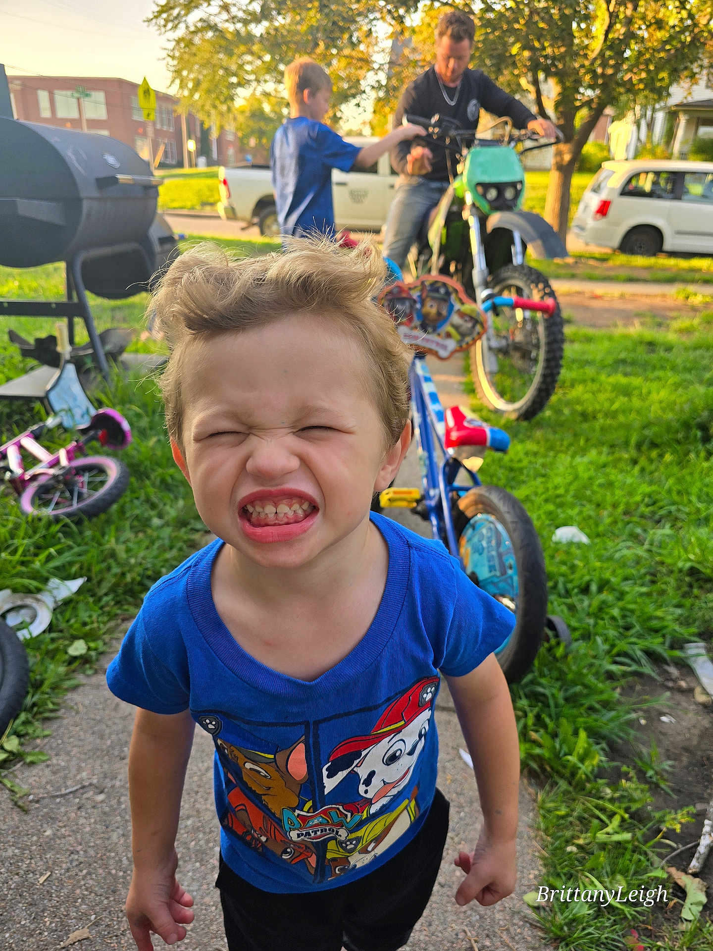 Thomas is registered to the contest to win money with this photo: child, boy, blue_shirt, bicycle, motorbike, outdoor, grass, sidewalk, playful, sunlight, person, toy, summer, parked_vehicle, residential_area, happy, scrunched_face, casual_clothing, daytime, urban