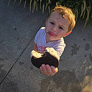 Thomas is registered to the contest to win money with this photo: child, toddler, cookie, food, hand, face, outdoor, concrete, grass, sunlight, messy, expression, offering, person, clothing, short_sleeve, footwear, sidewalk, nature, daylight