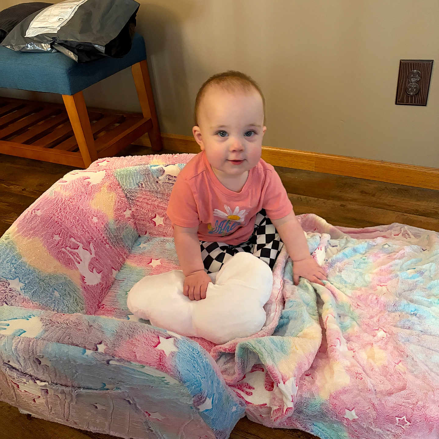 Olivia is registered to the contest to win money with this photo: baby, child, infant, blanket, pillow, heart_shaped_pillow, floor, wooden_floor, stool, furniture, pink, blue, plush, patterned_clothing, checkered_pants, toddler, indoor, smiling, cute, cozy