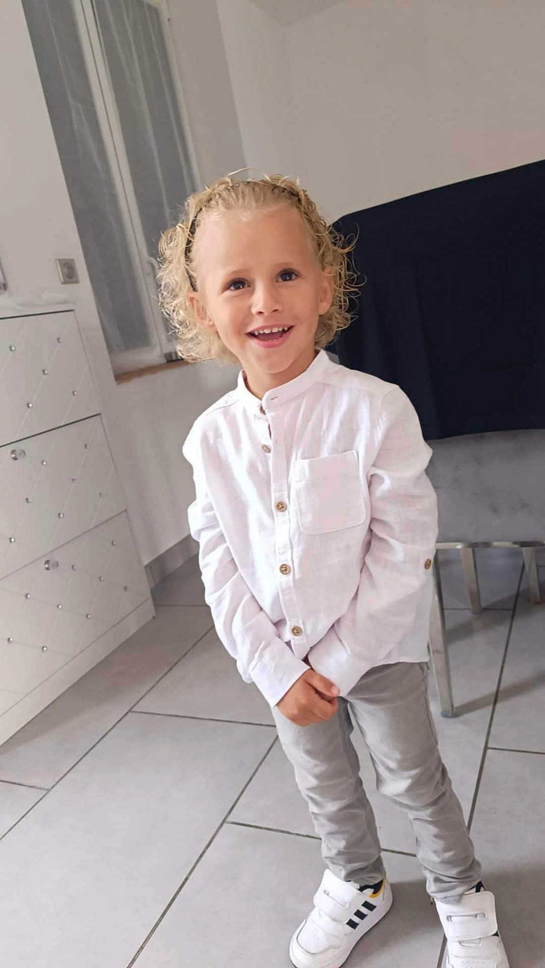 Tiago a rejoint le concours — aidez-le/la à gagner de superbes lots ! blond_hair, cabinet, casual_clothing, chair, child, curly_hair, grey_pants, happy, headband, indoor, light, person, portrait, smiling, standing, tile_floor, white_shirt, white_sneakers, window, young