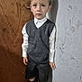 black_shoes, boy, casual_pose, child, curly_hair, earring, expression, fashion, floor, formal_clothing, gray_vest, indoor, light_shadow, portrait, shorts, standing, textured_wall, white_shirt, wooden_door, young_boy