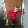 arms_outstretched, black_shoes, black_shorts, boy, casual_clothing, child, couch, door, door_handle, flooring, happy, home, indoors, light, playful, red_sweater, smile, wall, wooden_door, young_child