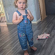 Everleigh is registered to the contest to win money with this photo: barefoot, blue_eyes, butterfly_embroidery, casual, child, clothing, curly_hair, denim_overalls, expression, flooring, hair_tuft, home, indoor, person, playroom, standing, toddler, toy, wooden_floor, young_child