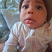 Xymir is registered to the contest to win money with this photo: baby, infant, child, portrait, close_up, face, wide_eyed, indoors, bed, bedding, knit_hat, hat, hoodie, jacket, clothing, zipper, sitting, soft_lighting, home, photography