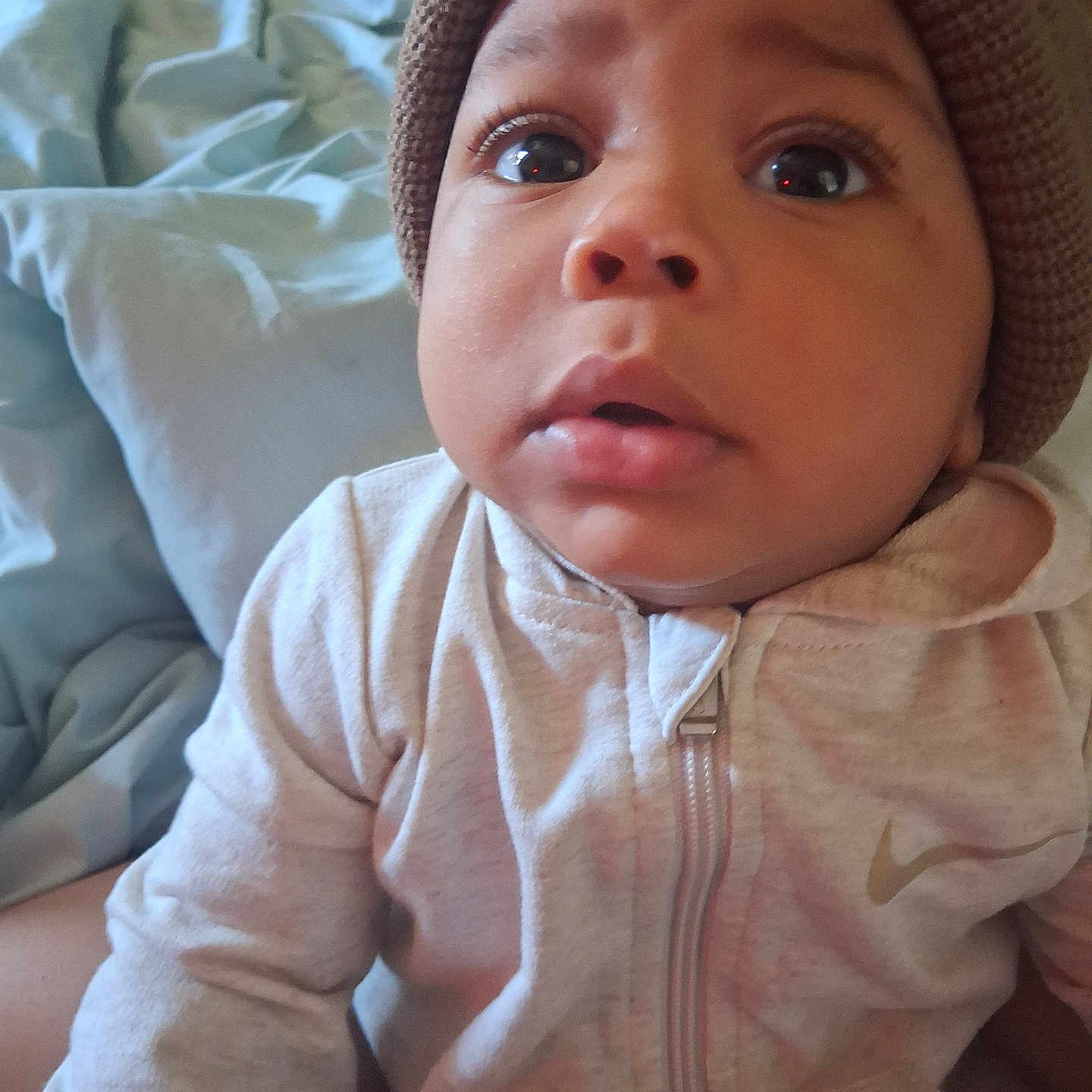 Xymir is registered to the contest to win money with this photo: baby, infant, child, portrait, close_up, face, wide_eyed, indoors, bed, bedding, knit_hat, hat, hoodie, jacket, clothing, zipper, sitting, soft_lighting, home, photography