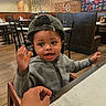 toddler, child, hat, cap, jacket, high_chair, hand, restaurant, table, wooden_floor, chair, person, indoor, casual, gesture, smiling, cute, dining, adult_hand, cozy