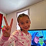 child, girl, smiling, peace_sign, braided_hair, pajamas, pink, indoors, television, people_on_tv, room, curtains, couch, chair, wall, ceiling, happy, portrait, casual, home
