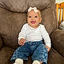 baby, child, smiling, headband, bow, jeans, heart_pattern, socks, white_shirt, armchair, brown_furniture, indoor, cute, happy, portrait, sitting, infant, clothing, person, cozy