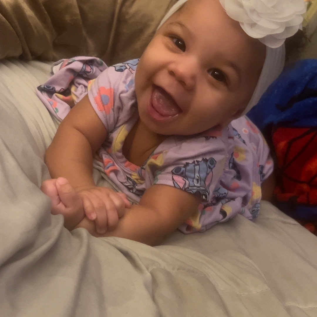 Zora is registered to the contest to win money with this photo: baby, smiling, headband, flower, bed, blanket, happy, infant, child, clothing, indoors, portrait, cute, toddler, hands, lying_down, person, soft_lighting, cozy, warm
