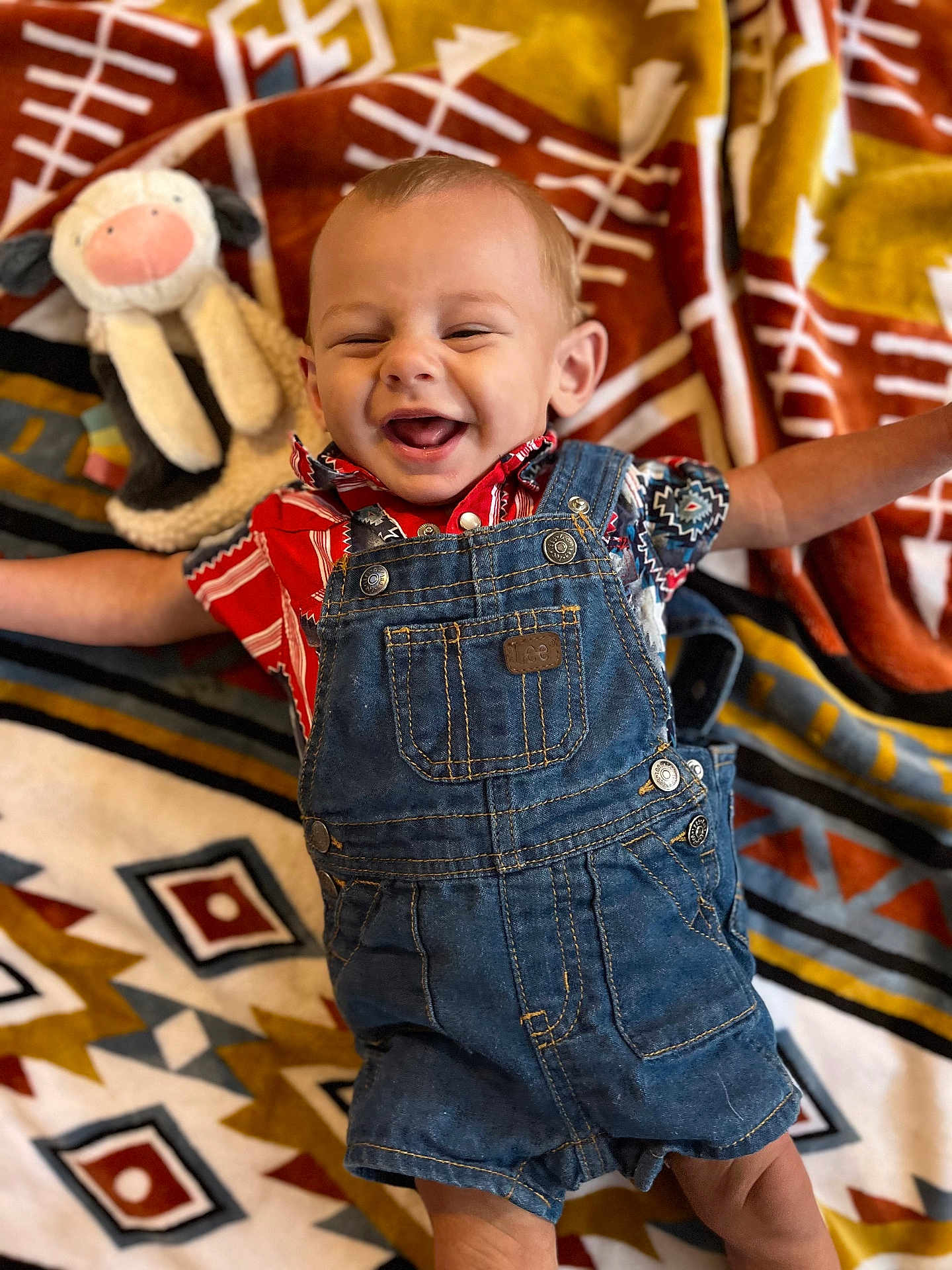 Hudson is registered to the contest to win money with this photo: baby, denim_overalls, smiling, stuffed_animal, colorful_clothing, blanket, patterned_blanket, happy, infant, cute, lying_down, child, toy, soft_toy, fabric, portrait, indoors, small_child, playful, joyful