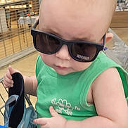 Thomas is registered to the contest to win money with this photo: baby, child, sunglasses, green_shirt, shopping_cart, indoor, person, holding, footwear, cute, fashion, head, face, casual, shopping, toddler, skin, hand, seat, retail