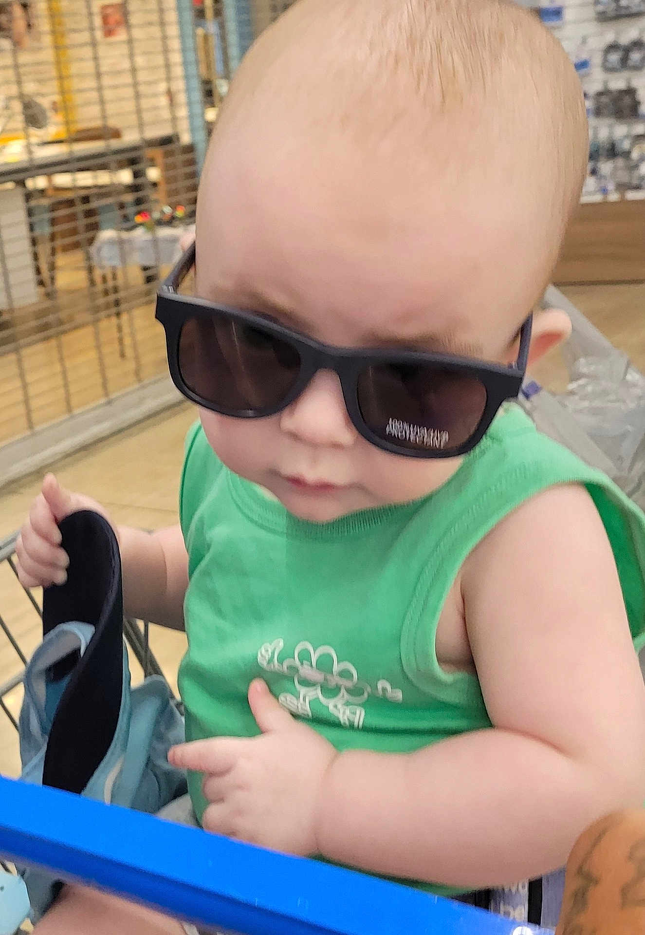 Thomas is registered to the contest to win money with this photo: baby, child, sunglasses, green_shirt, shopping_cart, indoor, person, holding, footwear, cute, fashion, head, face, casual, shopping, toddler, skin, hand, seat, retail