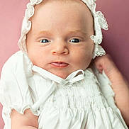 Annie is registered to the contest to win money with this photo: baby, infant, portrait, close_up, white_clothing, bonnet, curious_expression, soft_background, pink_background, newborn, cute, child, face, skin, headwear, young, person, indoors, smile, small_hands