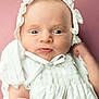 baby, infant, portrait, close_up, white_clothing, bonnet, curious_expression, soft_background, pink_background, newborn, cute, child, face, skin, headwear, young, person, indoors, smile, small_hands