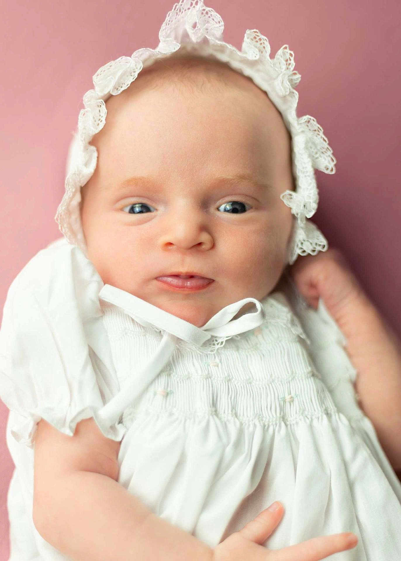 Annie is registered to the contest to win money with this photo: baby, infant, portrait, close_up, white_clothing, bonnet, curious_expression, soft_background, pink_background, newborn, cute, child, face, skin, headwear, young, person, indoors, smile, small_hands