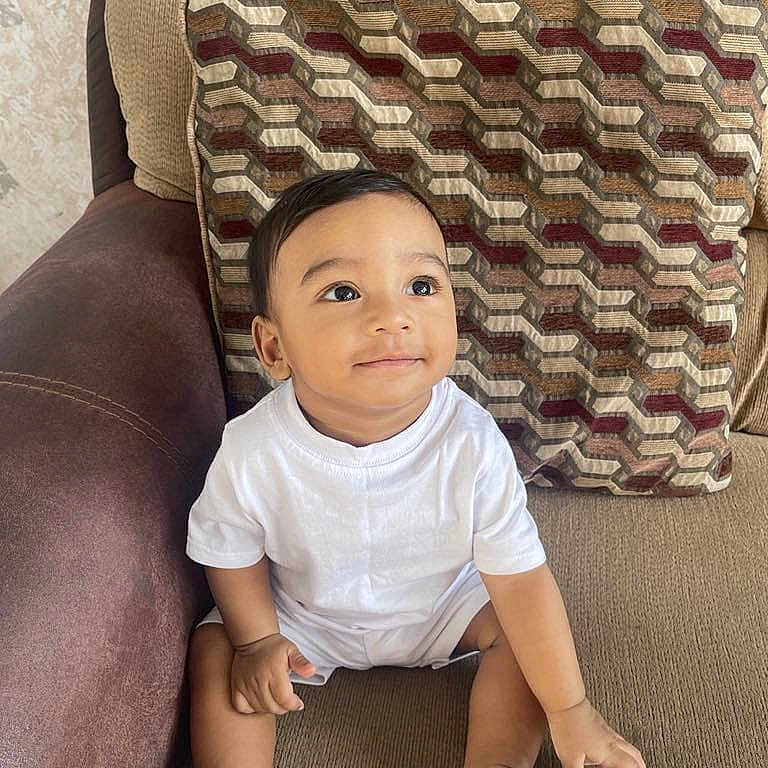 Ahil joined the competition — help win amazing prizes! toddler, child, sitting, white_clothing, sneakers, couch, cushion, pattern, indoor, young_child, small_shoes, curious_expression, person, furniture, home, face, looking_up, cute, casual, relaxed