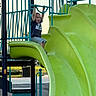 child, playground, slide, play_equipment, outdoor, park, green, blue, clothing, shorts, tshirt, tongue_out, fun, happy, summer, daylight, metal, plastic, bars, recreation
