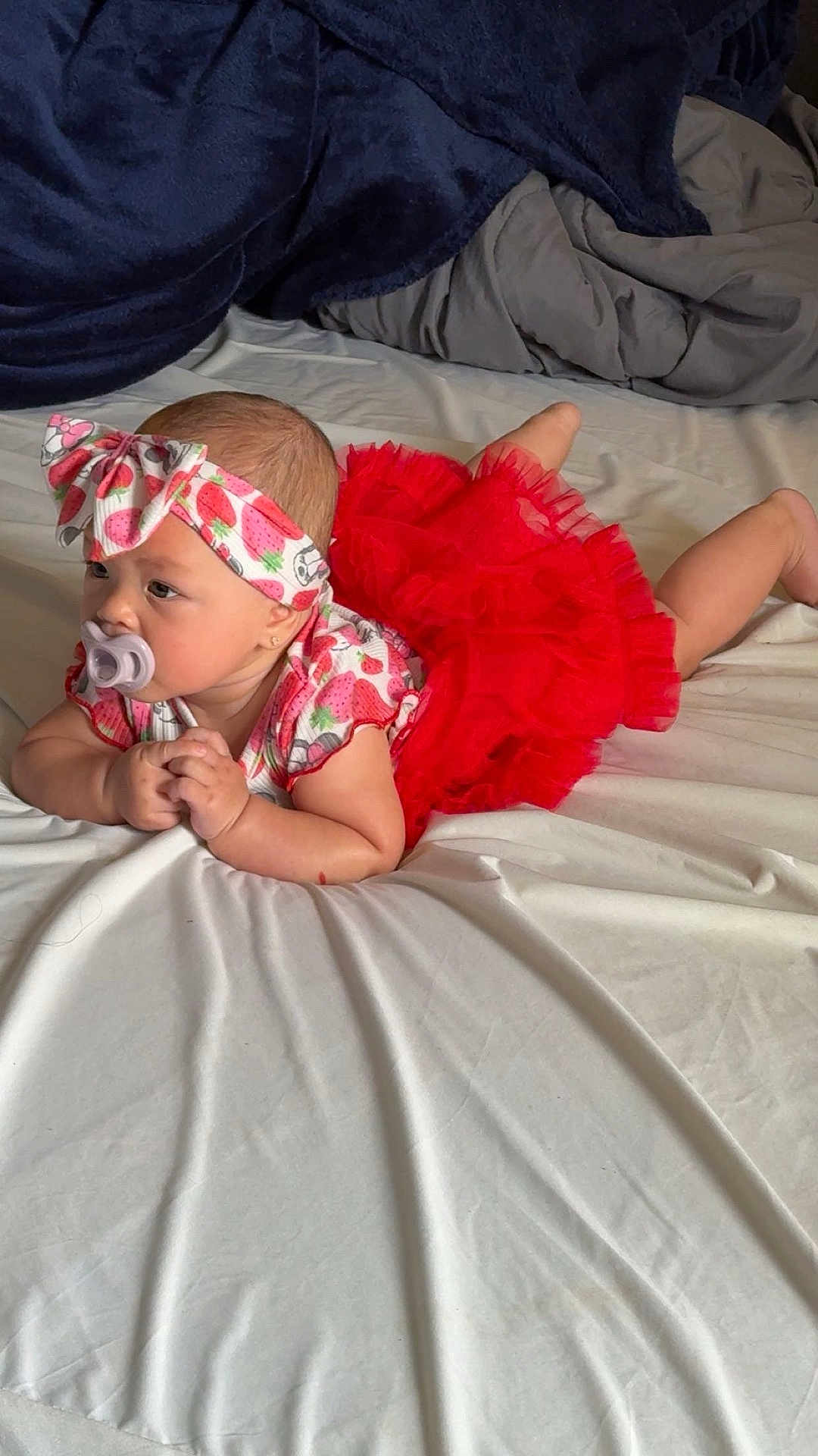 Sabrina is registered to the contest to win money with this photo: baby, infant, pacifier, bed, blanket, red_skirt, headband, strawberry_pattern, clothing, cute, lying_down, indoor, person, skin, arms, expression, resting, toddler, soft_fabric, comfort