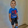 child, boy, smiling, standing, blue_tshirt, spiderman, shorts, sneakers, blue_shoes, white_socks, wooden_floor, wall, indoor, casual_clothing, happy, portrait, person, short_hair, watch, flooring