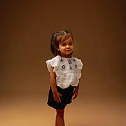 Alya participe au concours pour gagner de l'argent avec cette photo : child, girl, barefoot, white_top, embroidered_clothing, shorts, standing, smiling, posing, brown_background, studio_lighting, portrait, cute, young, fashion, hair_tied, innocent, playful, full_body, casual