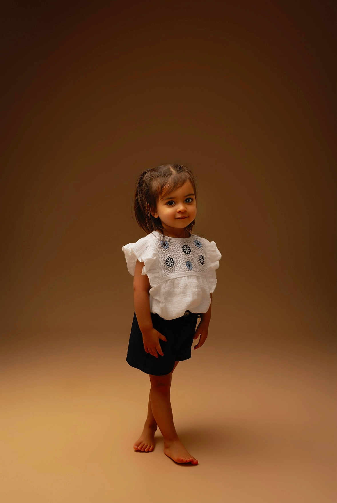 Alya participe au concours pour gagner de l'argent avec cette photo : child, girl, barefoot, white_top, embroidered_clothing, shorts, standing, smiling, posing, brown_background, studio_lighting, portrait, cute, young, fashion, hair_tied, innocent, playful, full_body, casual