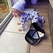 Adonis is registered to the contest to win money with this photo: baby, child, crawling, onesie, hat, pom_pom, black_cap, floor, wooden_floor, tile_floor, glass_door, indoor, curious, happy, smiling, face, hand, footwear, clothing, person