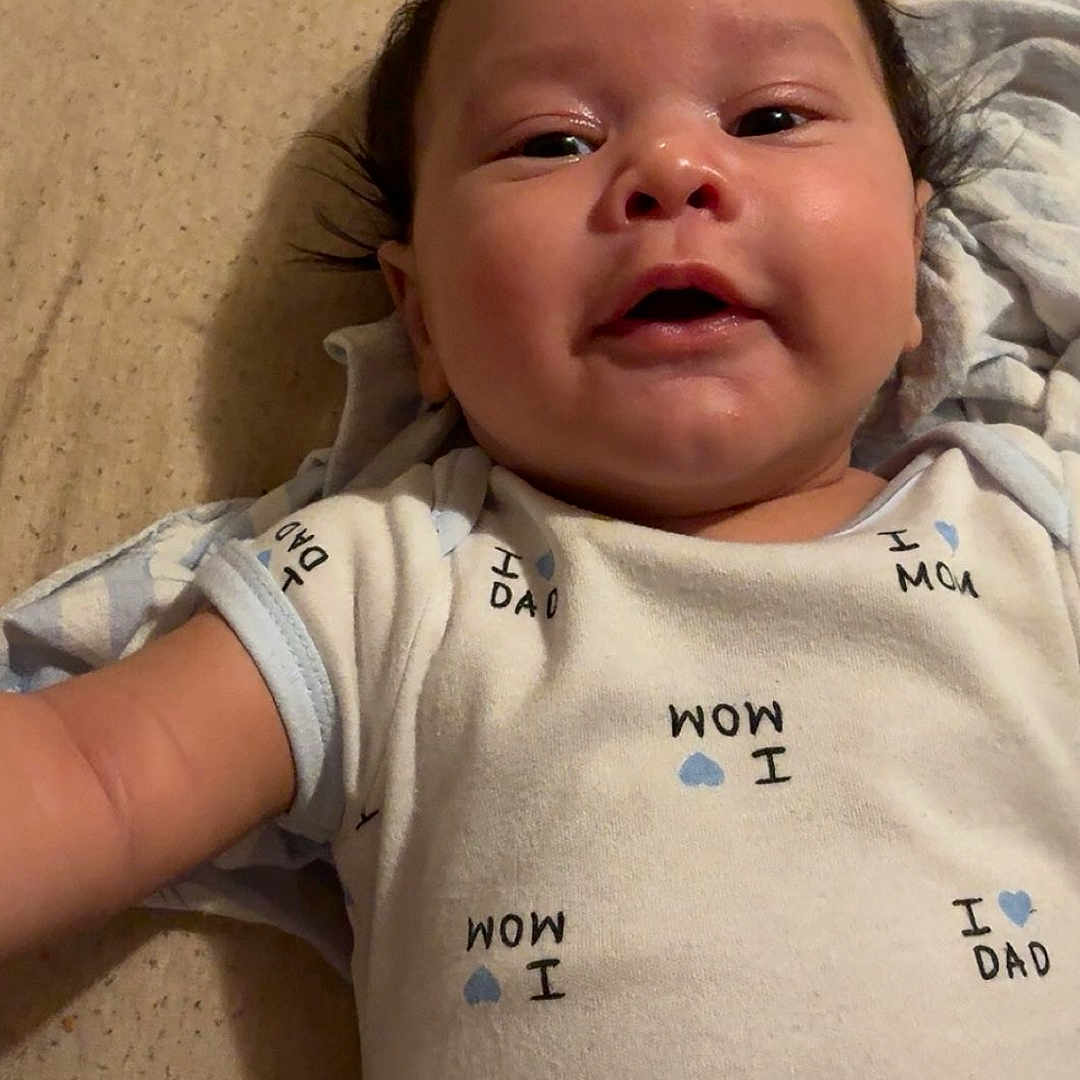 Swayde is registered to the contest to win money with this photo: arm, baby, bed, blanket, child, clothing, cute, face, happy, head, indoors, infant, lying_down, newborn, onesie, person, portrait, skin, smiling, young_child