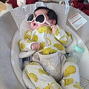 Celeste is registered to the contest to win money with this photo: baby, baby_rocker, sunglasses, heart_shaped, onesie, green_apple_pattern, soft_toy, bottle, infant, cozy, indoor, cute, smiling, baby_clothes, plush_toy, seat, relaxed, child, playful, comfort