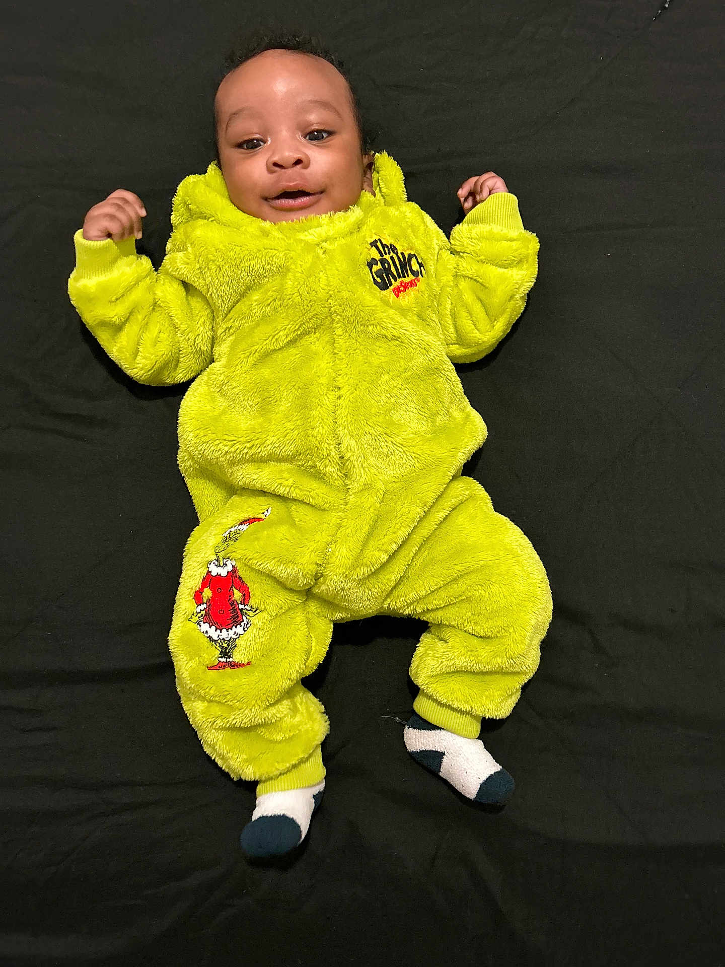 Brooklyn is registered to the contest to win money with this photo: baby, child, smile, green_onesie, grinch, socks, lying_down, black_background, infant, cute, happy, cozy_clothing, portrait, baby_clothes, childrens_wear, indoors, one_person, young_child, bright_color, soft_texture