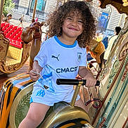 Maïronn a rejoint le concours — aidez-le/la à gagner de superbes lots ! child, smiling, curly_hair, carousel, amusement_ride, tiger_figure, handlebars, white_shirt, blue_trim, shorts, outdoor, daylight, playful, fun, happy, person, seat, background_people, urban, joy