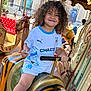 child, smiling, curly_hair, carousel, amusement_ride, tiger_figure, handlebars, white_shirt, blue_trim, shorts, outdoor, daylight, playful, fun, happy, person, seat, background_people, urban, joy