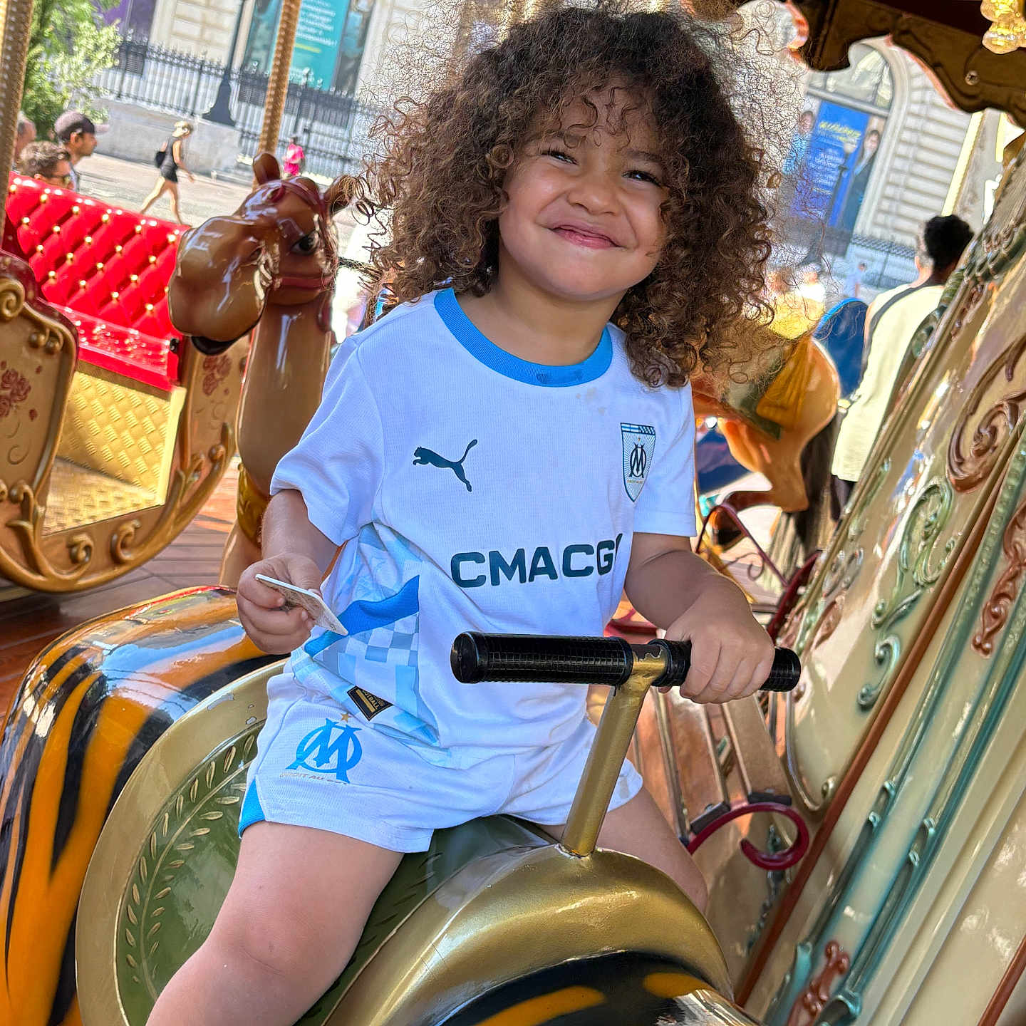 Maïronn a rejoint le concours — aidez-le/la à gagner de superbes lots ! amusement_ride, background_people, blue_trim, carousel, child, curly_hair, daylight, fun, handlebars, happy, joy, outdoor, person, playful, seat, shorts, smiling, tiger_figure, urban, white_shirt