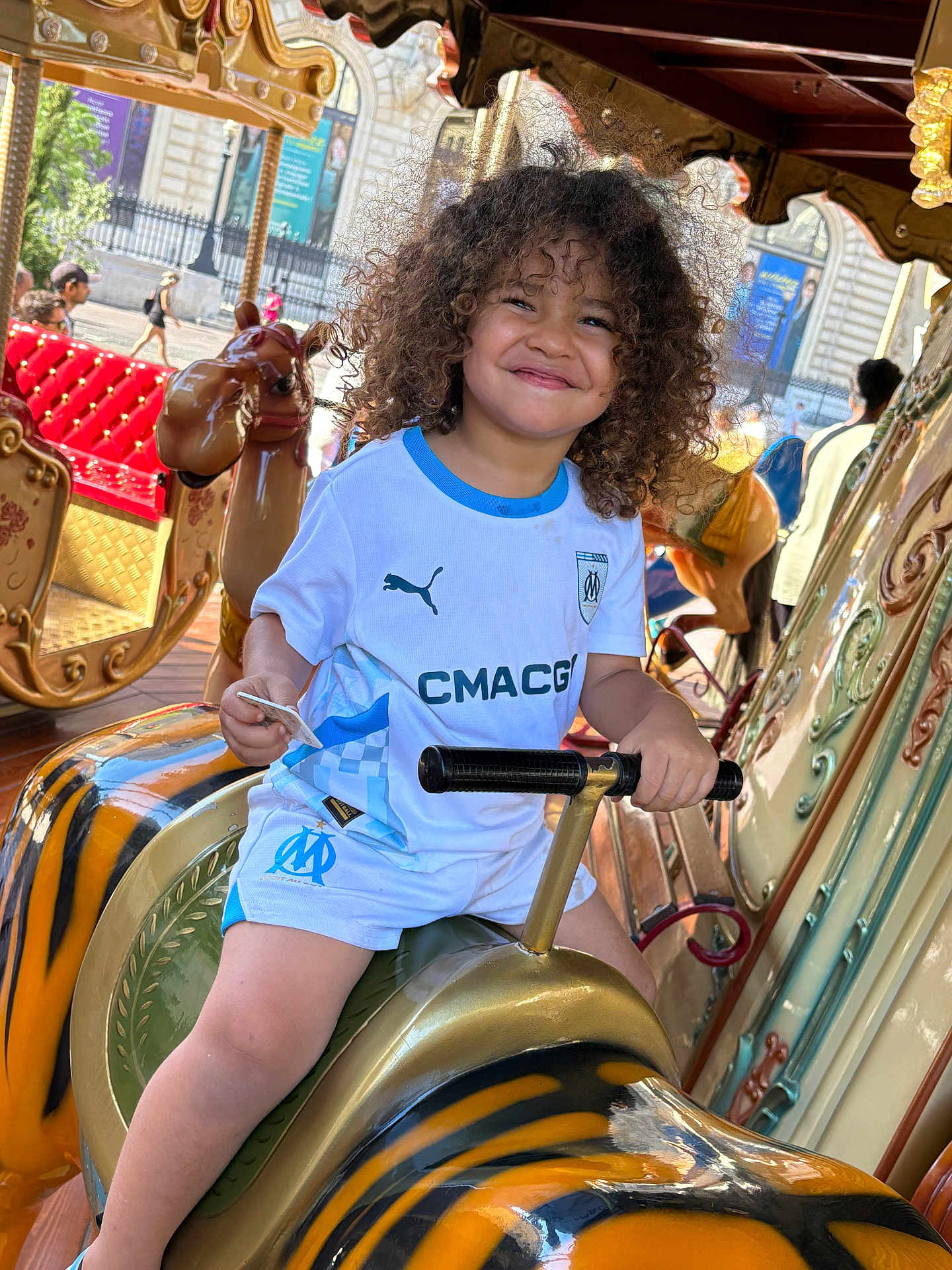 Maïronn a rejoint le concours — aidez-le/la à gagner de superbes lots ! child, smiling, curly_hair, carousel, amusement_ride, tiger_figure, handlebars, white_shirt, blue_trim, shorts, outdoor, daylight, playful, fun, happy, person, seat, background_people, urban, joy