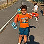 child, boy, scooter, outdoor, smiling, orange_shirt, blue_shorts, white_shoes, sunlight, paved_path, shadow, curb, fence, grass, trees, people, summer, play, happy, casual