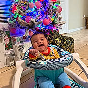 Neithan is registered to the contest to win money with this photo: baby, baby_seat, child, christmas_tree, clothing, decorations, festive, floor, happy, holiday, home, indoor, lights, ornaments, patterned, pointing, presents, smiling, socks, tiled_floor