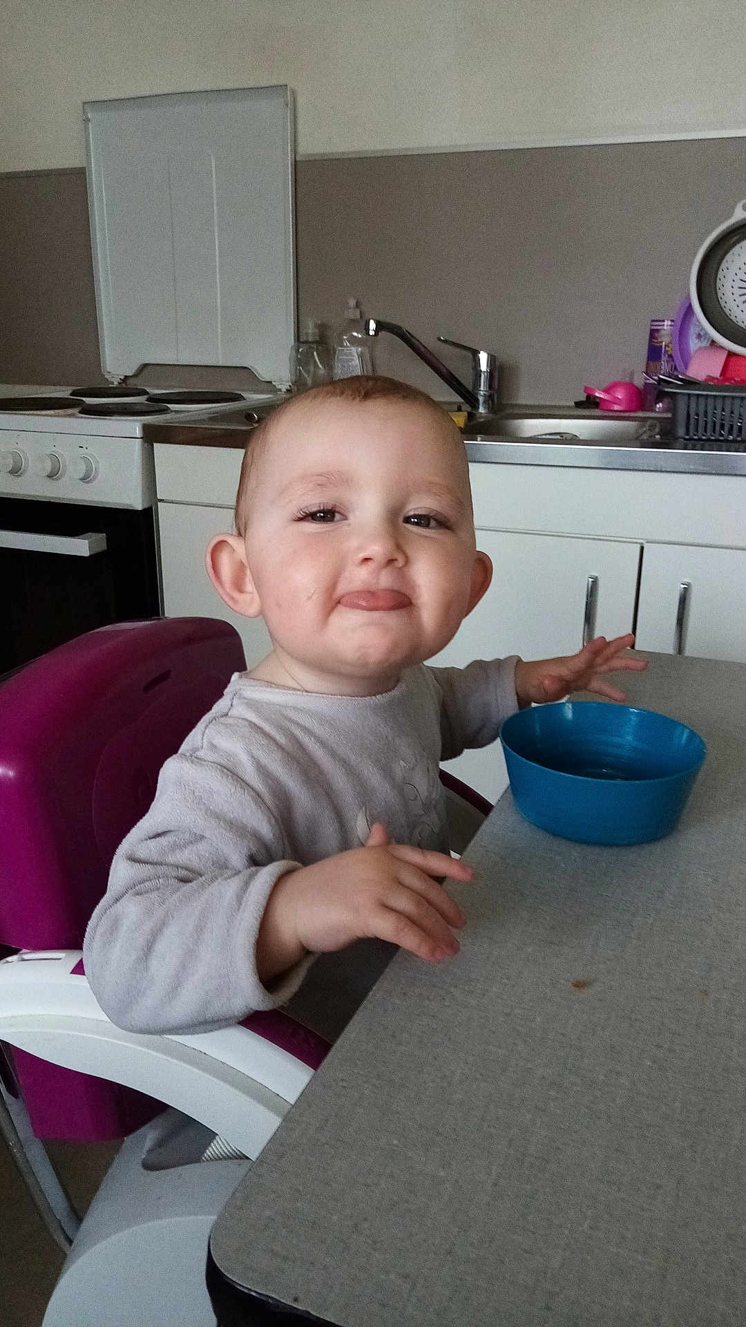 Alya a rejoint le concours — aidez-le/la à gagner de superbes lots ! child, toddler, highchair, bowl, kitchen, table, face, smile, tongue_out, ear, eyes, stove, sink, faucet, cabinet, cupboard, plastic_bowl, casual_clothing, mealtime, portrait