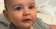 Adam is registered to the contest to win money with this photo: baby, child, infant, portrait, close_up, face, eyes, wide_eyed, curious, looking_at_camera, short_hair, gray_shirt, clothing, sitting, bed, bedding, indoor, home, soft_lighting, photograph