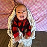Corey is registered to the contest to win money with this photo: baby, blanket, checkered_pattern, red_clothing, black_clothing, smiling, happy, infant, couch, pillow, pink_pillow, cozy, wrapped, indoor, portrait, cute, child, seated, home, soft